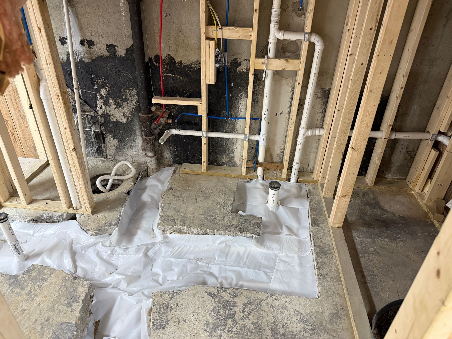 Basement bathroom construction: wood framing, plumbing pipes, concrete floor with debris and protective fabric.