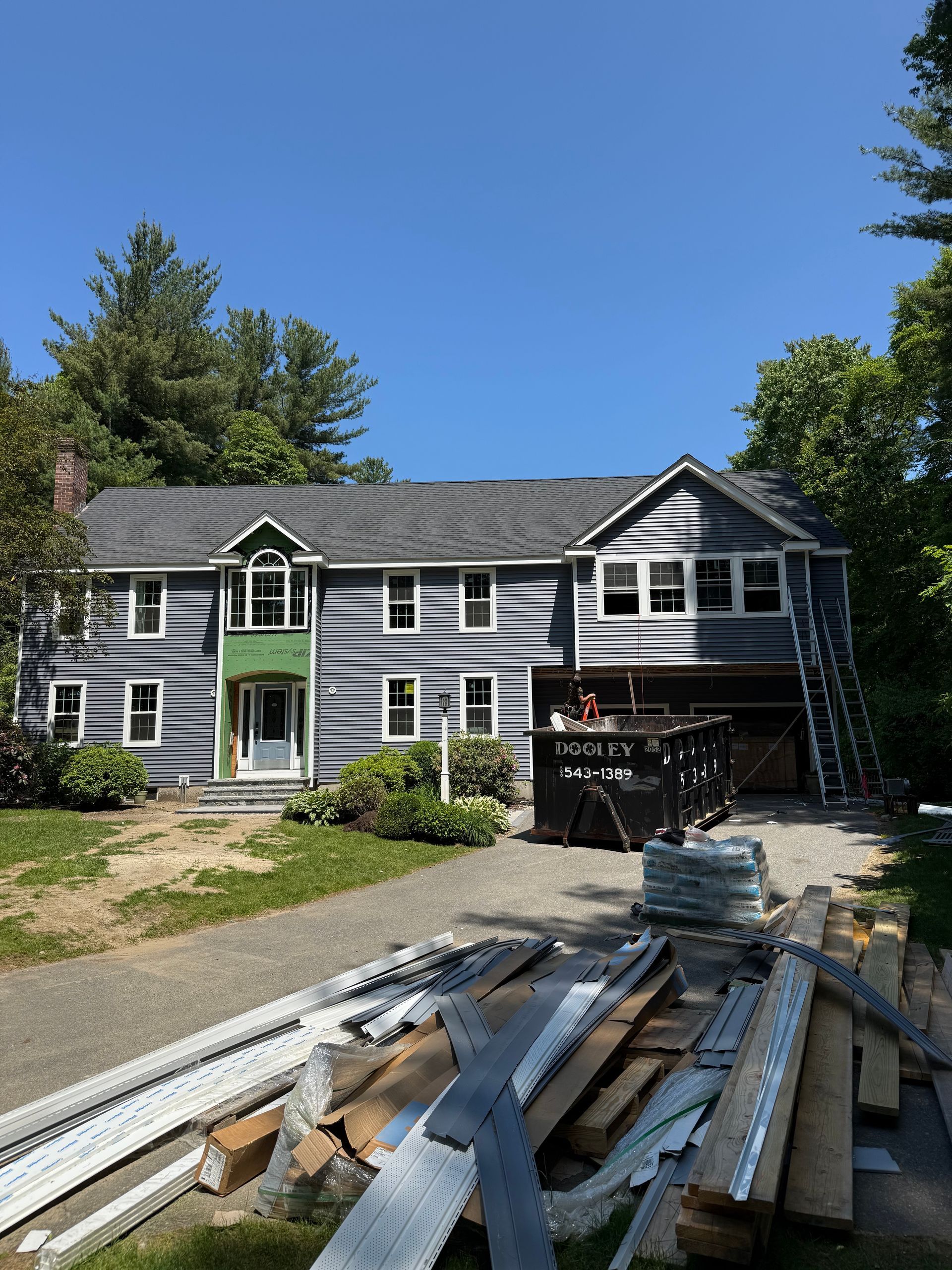 House under construction, grey siding, white trim, with materials and a dumpster in front, sunny day.