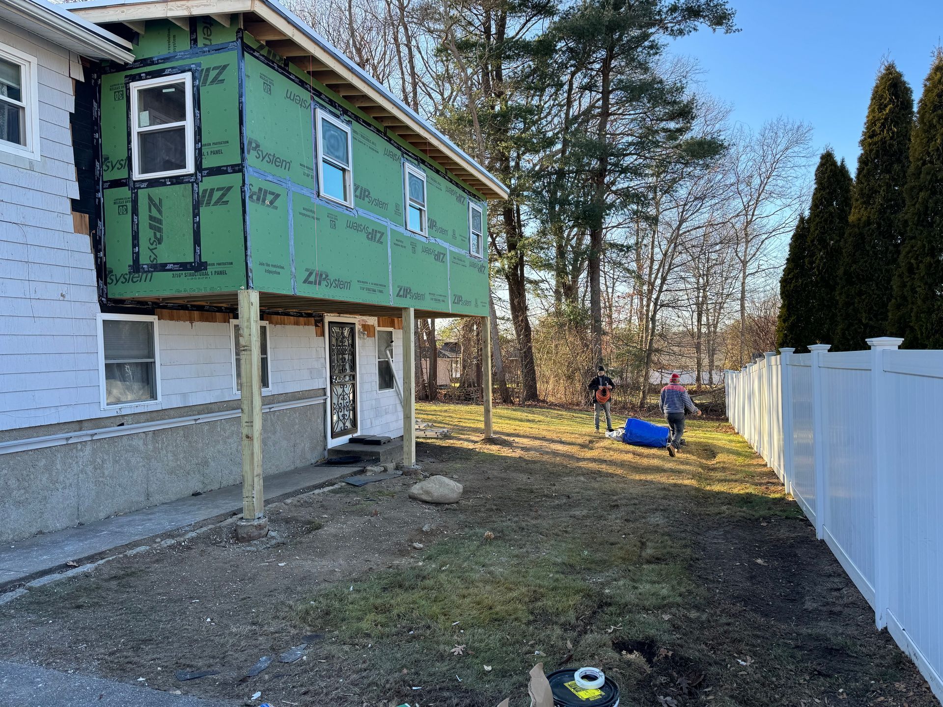 Exterior house under construction with elevated addition on wooden posts. Two people in yard, white fence.