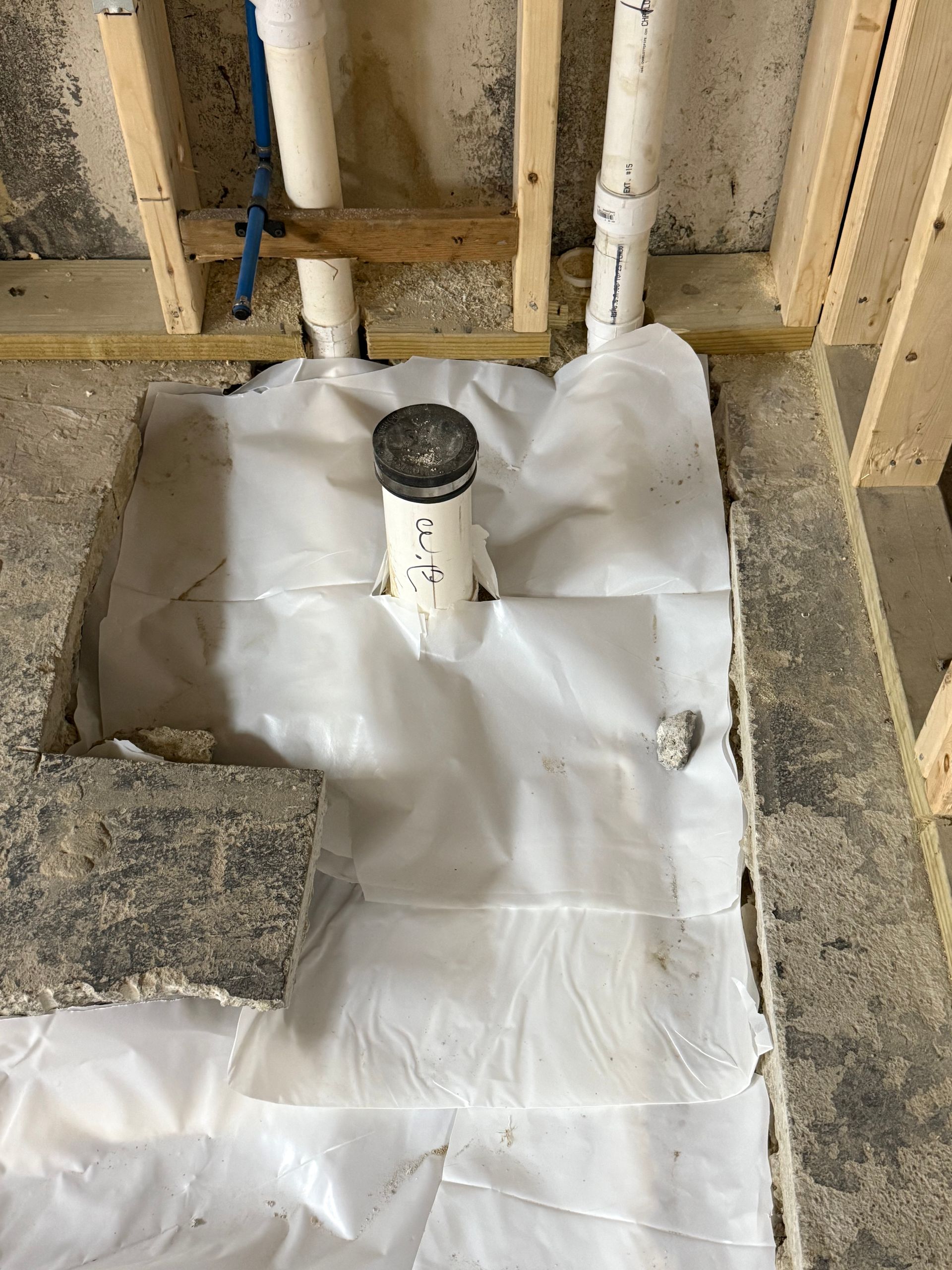 Framed-out bathroom floor with plumbing: drain pipe, plastic sheeting, unfinished concrete, and wooden framing.