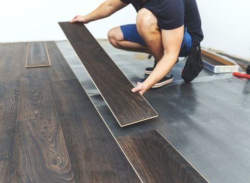 Person installing dark wood laminate flooring, kneeling on a concrete surface.