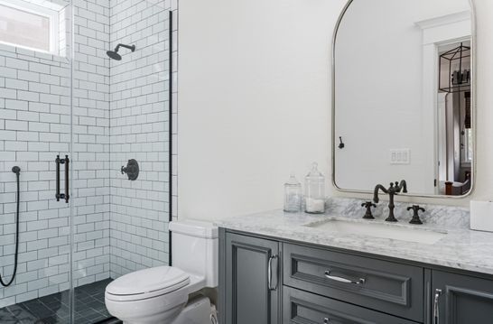 Bathroom with white subway tile shower, gray vanity, toilet, and arched mirror.