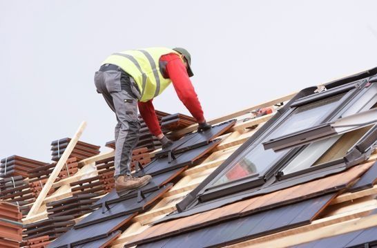 Roofer in safety vest laying tiles on a rooftop with skylights, cloudy day.