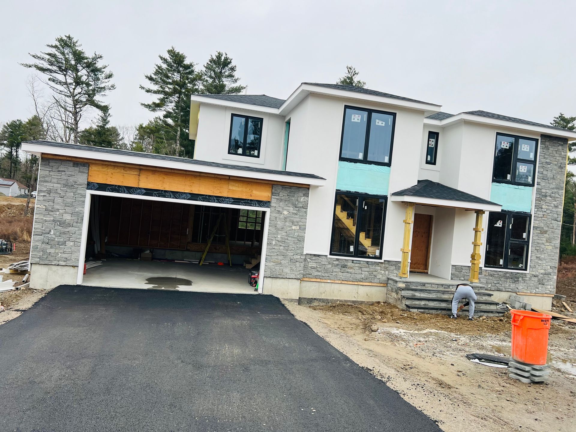 Modern house under construction with a black asphalt driveway and stone accents. A worker is visible.