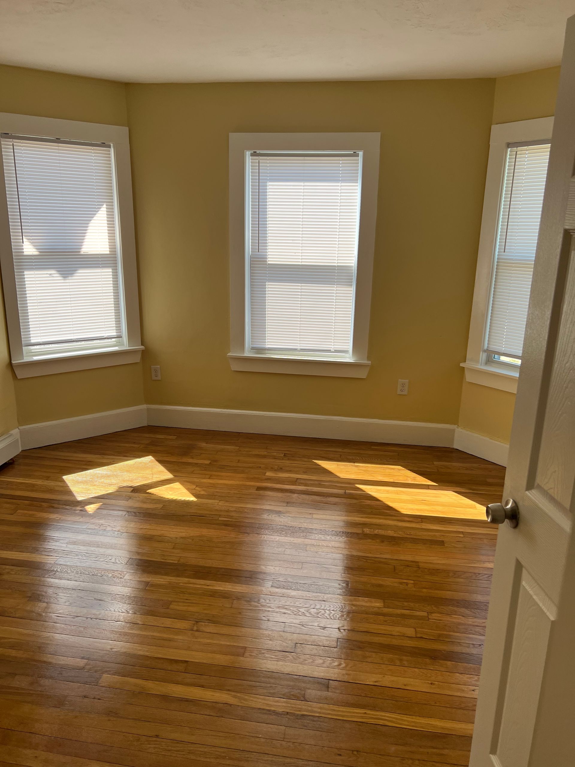 Empty room with yellow walls, hardwood floor, and three windows with blinds.