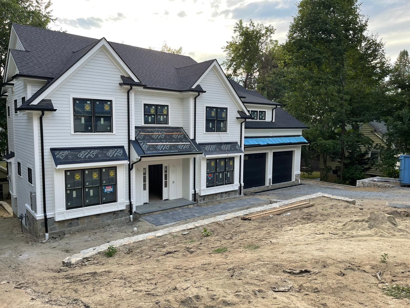 White two-story house with black trim, unpaved driveway, under construction; set on a hillside.