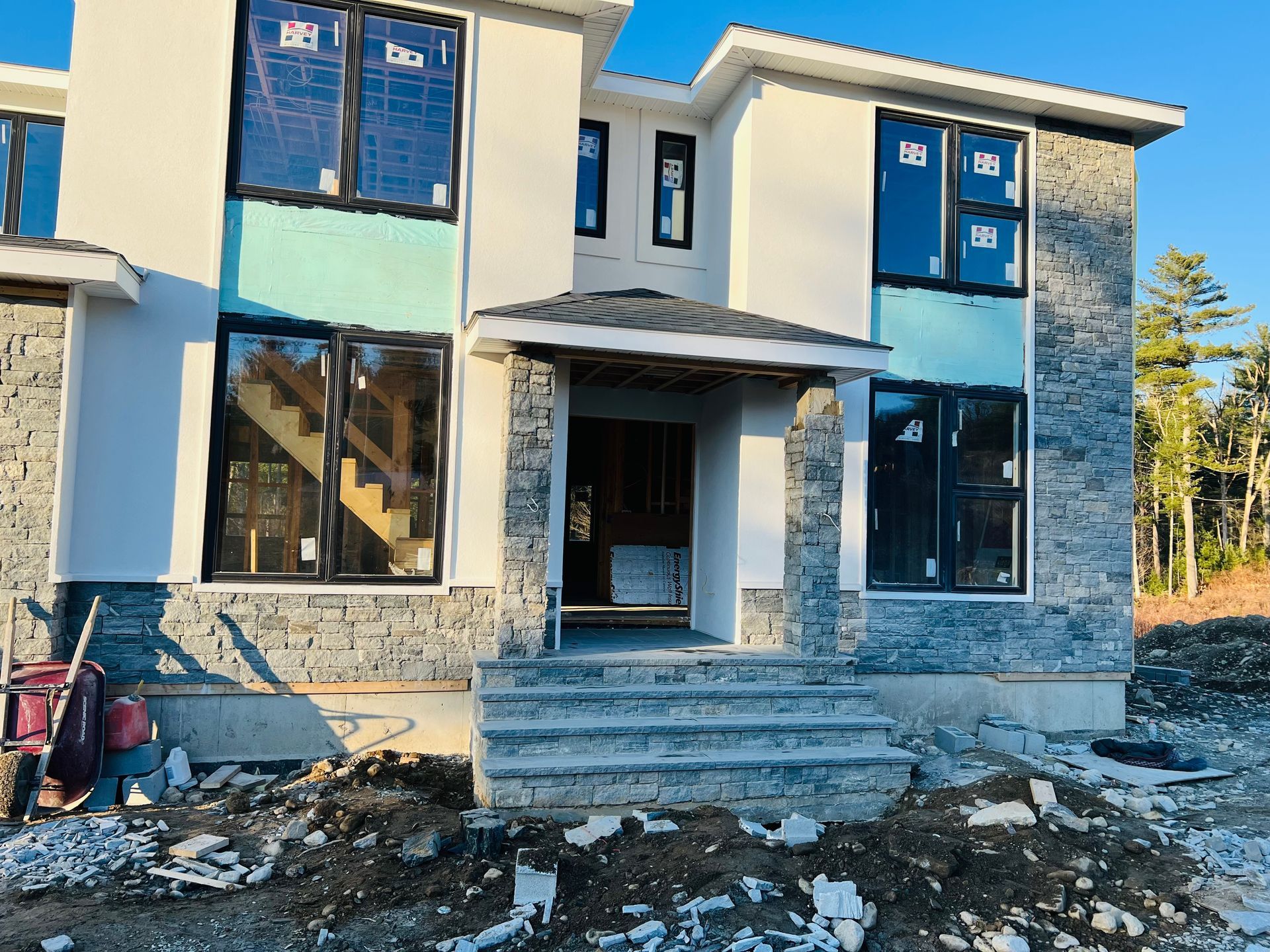 Two-story house under construction with gray stone facade, large black-framed windows, and light blue accents.