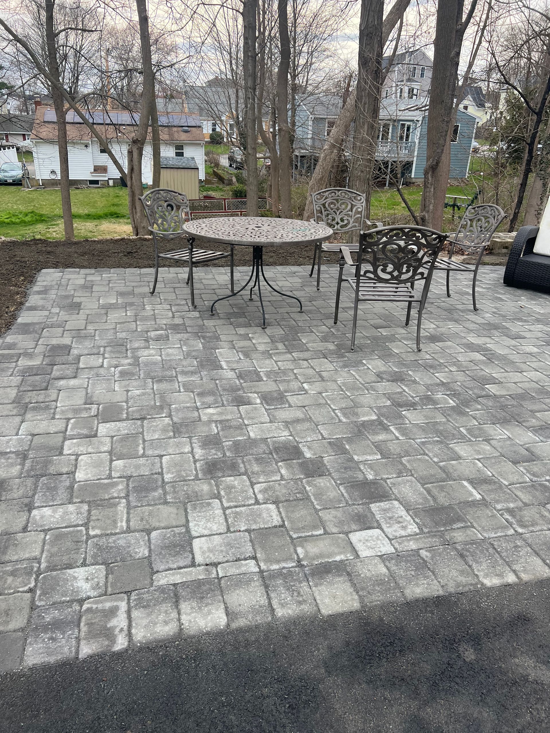 Patio paved with gray bricks, with ornate metal table and chairs, near trees and houses.