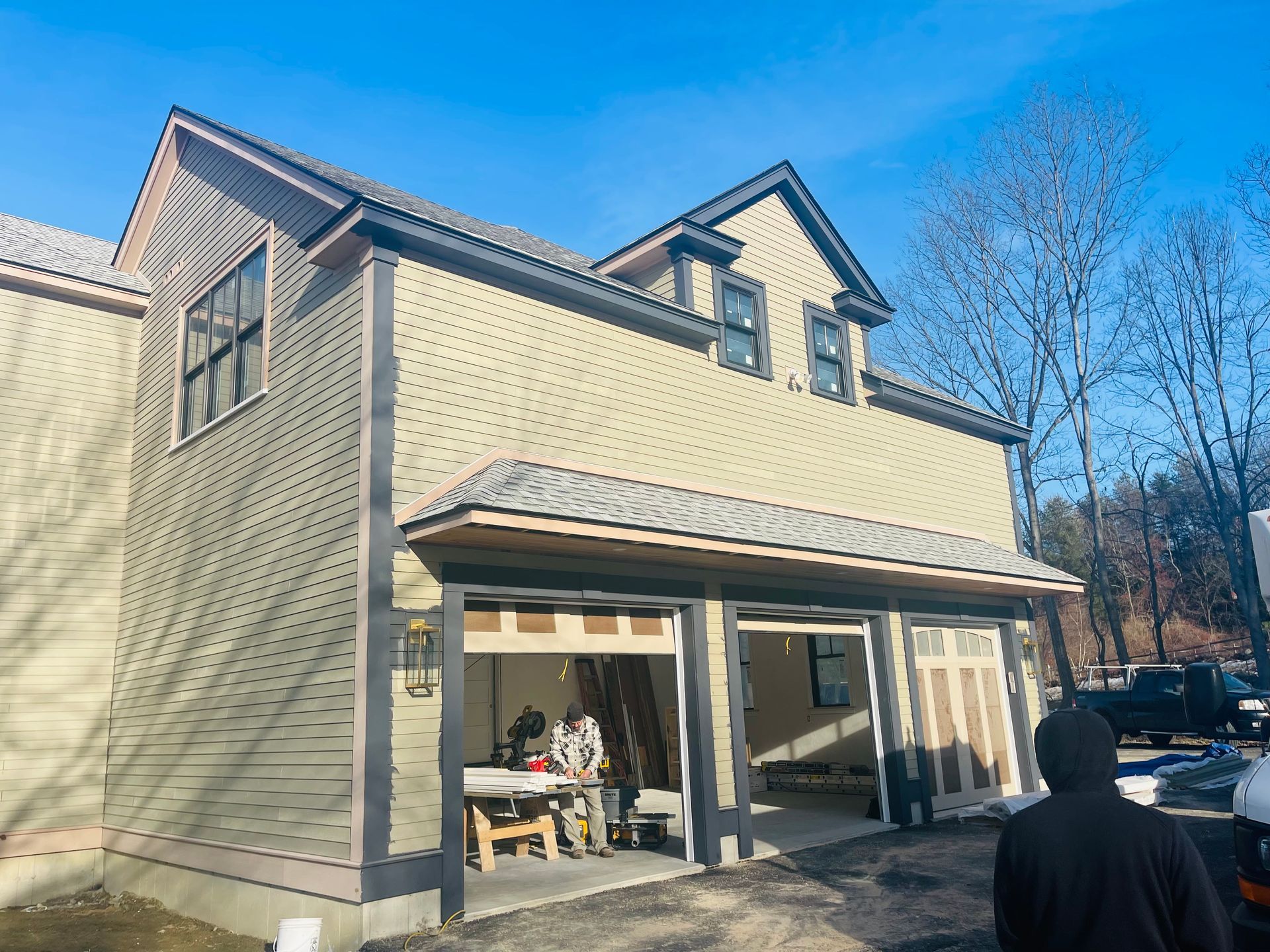 Two-story building with garage doors open, two people inside, tan siding, grey trim, blue sky.