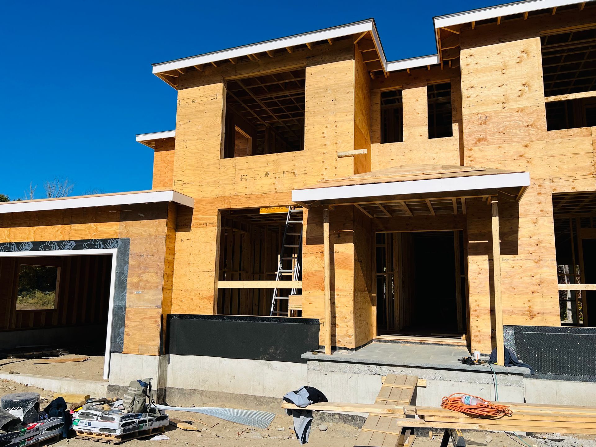 Construction of a two-story house, wooden frame exposed. Entry porch, garage, and windows are visible against a blue sky.