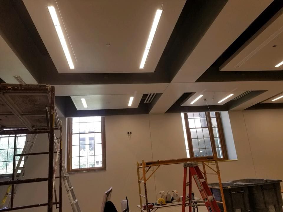 Interior of a room under construction with lit ceiling panels, scaffolding, windows, and ladders.