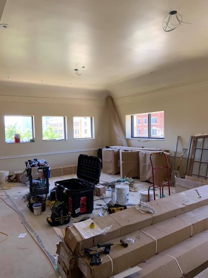 Interior renovation with tools, materials, and covered objects. Light-colored walls and large windows in room.