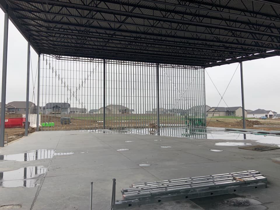 Construction site with metal framing, concrete floor, and open roof. Houses in the background.