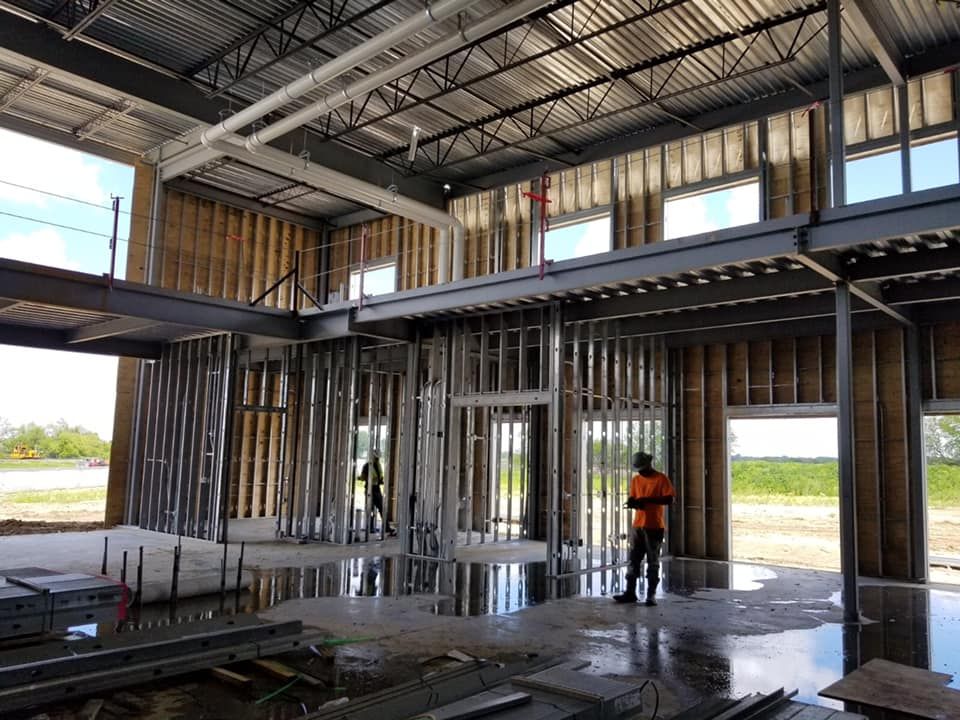 Construction site interior: metal framing, concrete floor, workers. Windows and a mezzanine level are visible.