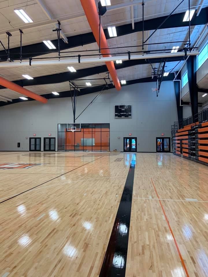 Basketball court interior, wooden floor with black lines, orange and black accents, bleachers, scoreboard.