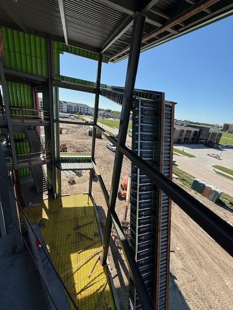 Construction site interior: Steel beams, yellow flooring, green insulation, and outdoor view on a sunny day.