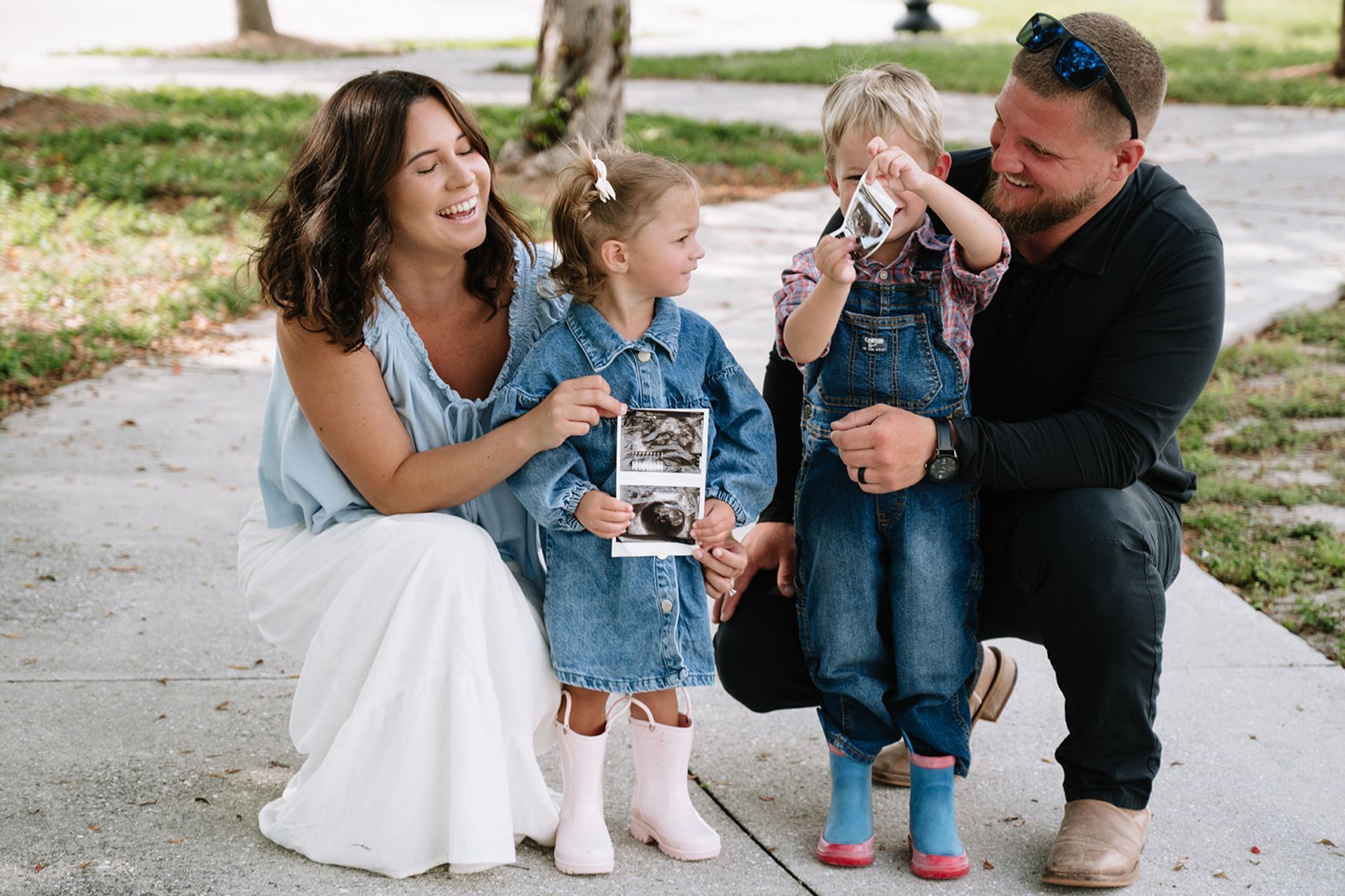 Family seated on a sidewalk, smiling and holding two children in denim outfits outdoors