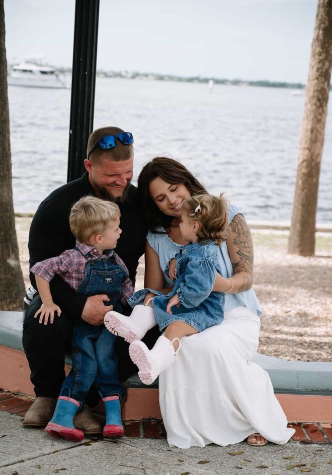 Family sitting on a bench by the water, smiling and cuddling two small children