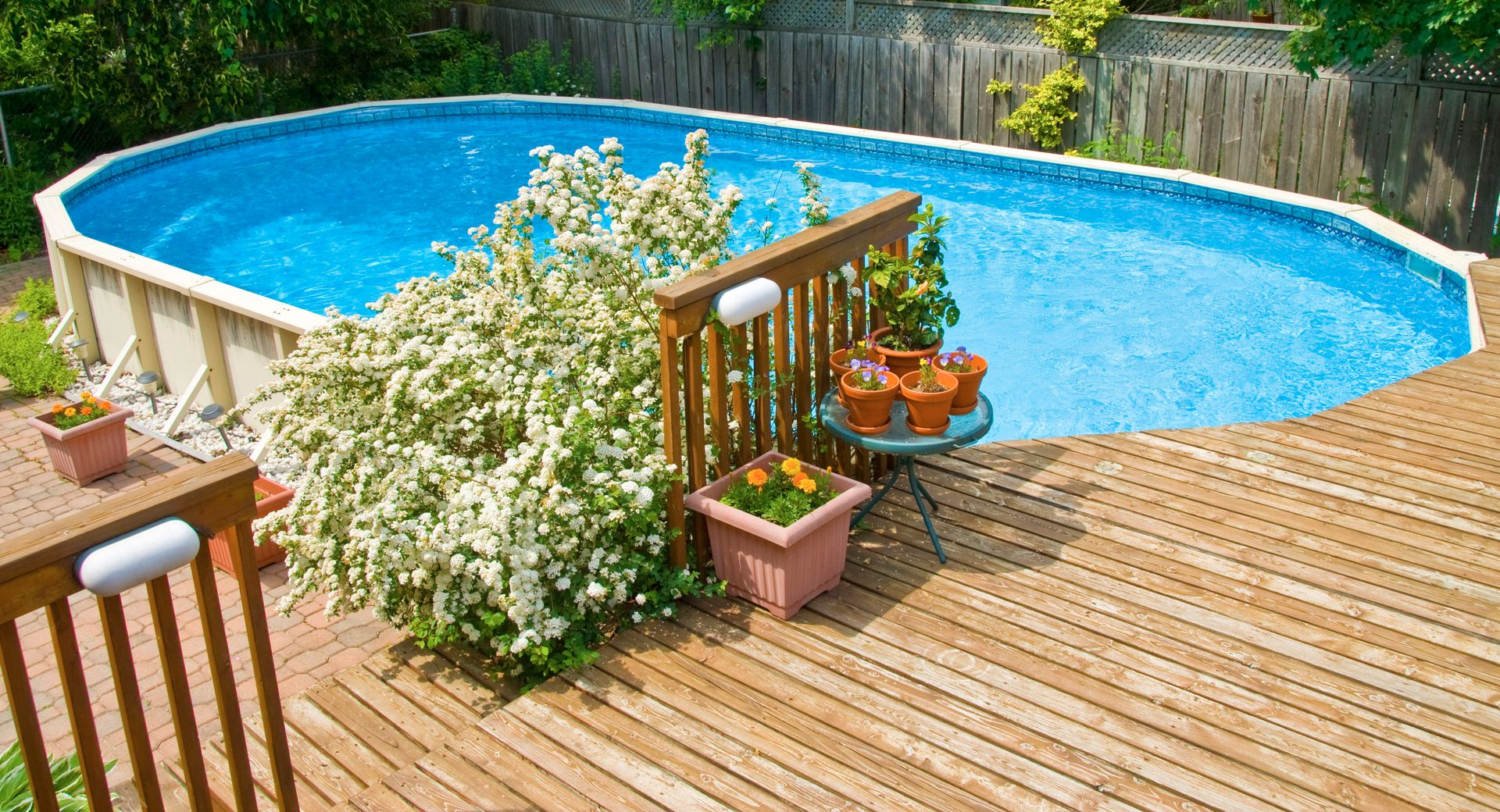 Backyard pool with blue water beside a wooden deck and potted flowers near a fence