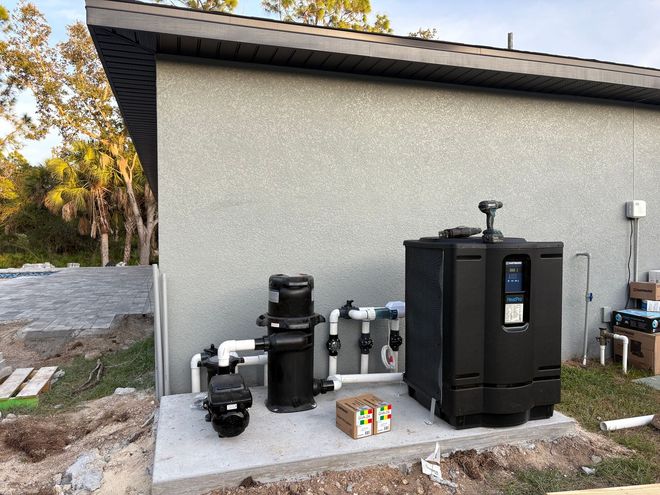 Outdoor pool equipment beside a gray house, with black pumps, filters, and heater units on a concrete pad