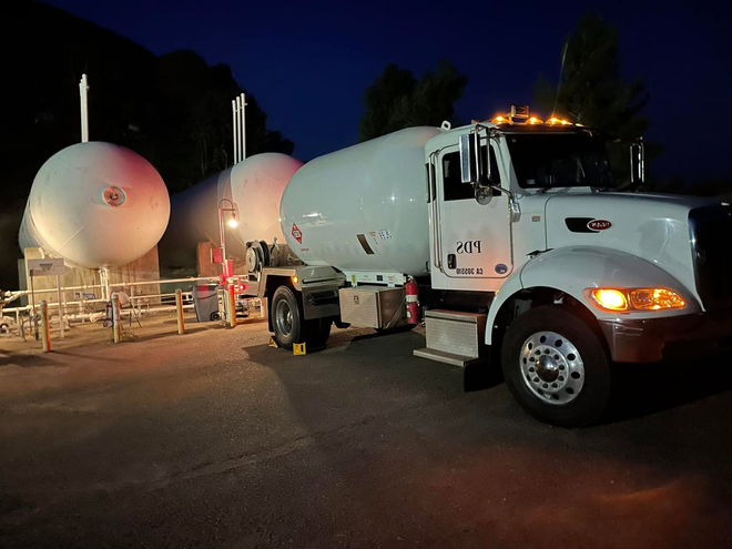 White tanker truck parked beside large round tanks at night, lit by work lights.
