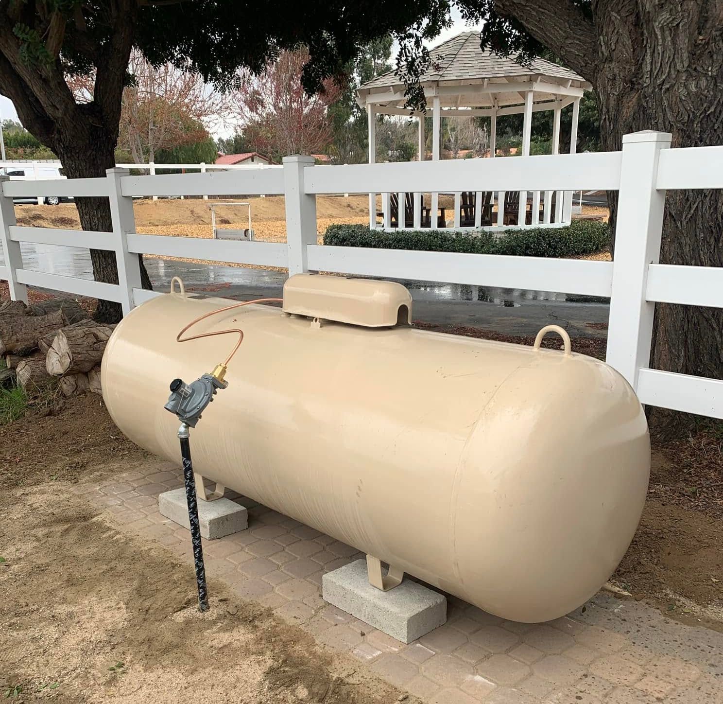 Beige propane tank beside a white fence in a yard, with a valve and small utility cover.