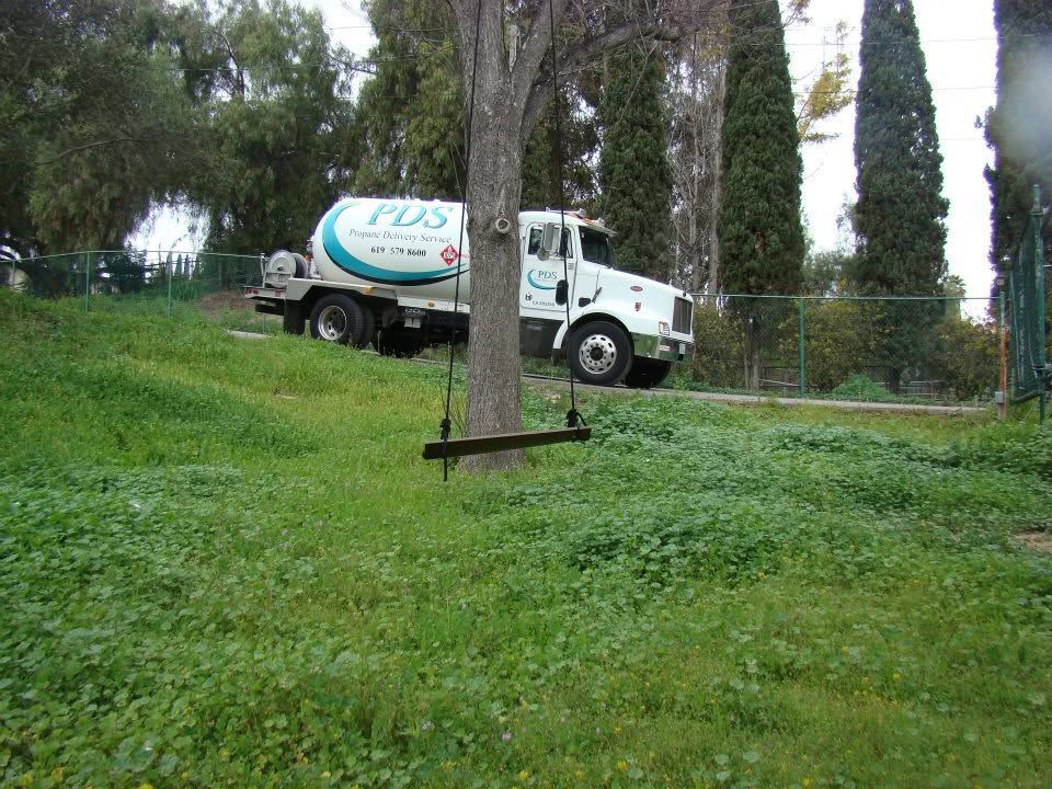 White tanker truck parked on a grassy slope by trees and a fence