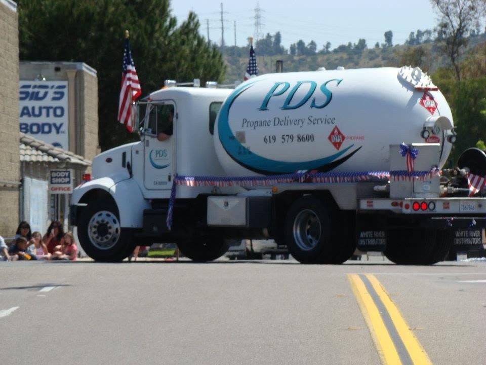 White PDS fuel truck crossing a street in a parade, with spectators and an American flag nearby