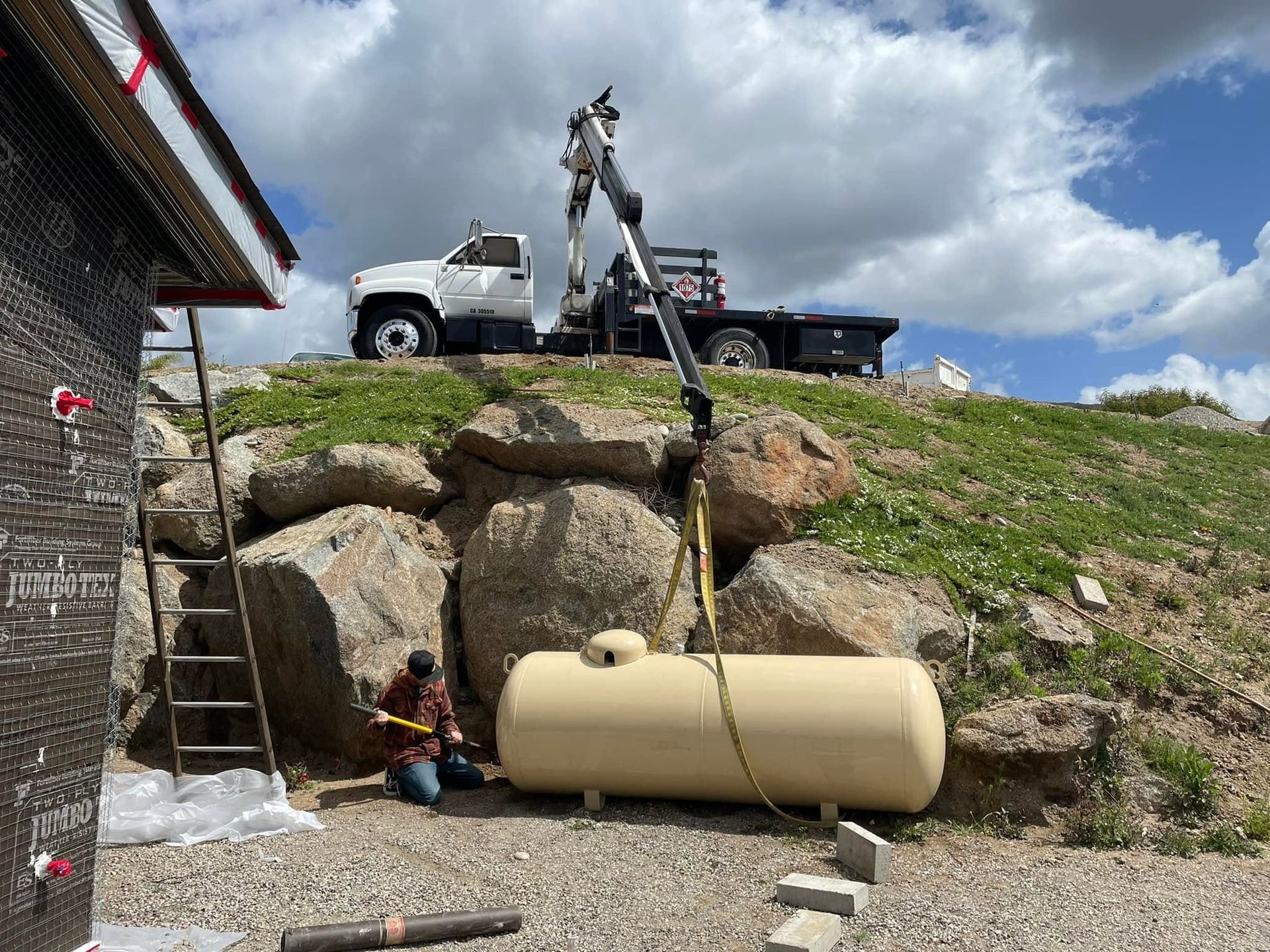 Worker operating boom lift above large beige tank by a rocky hillside and building