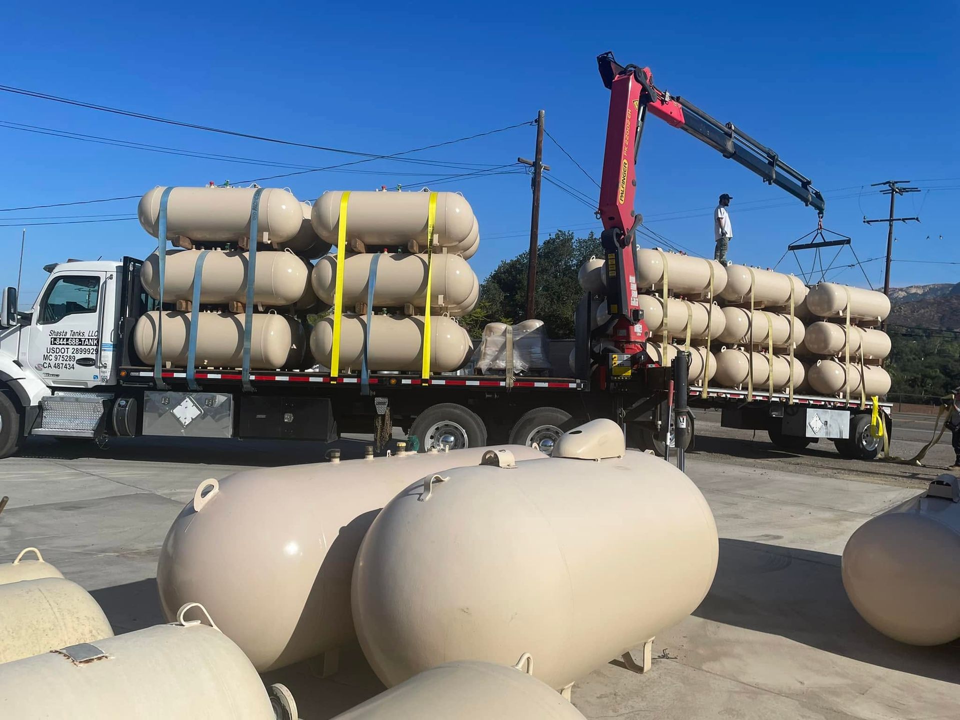 Flatbed trucks loaded with tan cylindrical tanks at an outdoor work site under a blue sky