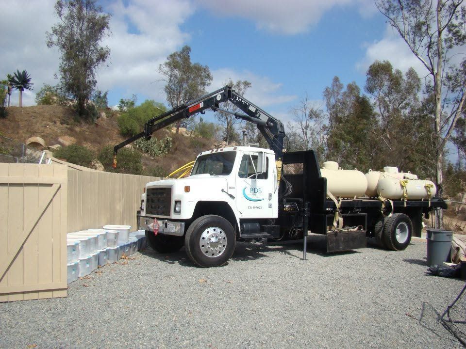White utility truck with crane beside beige fence and tanks on a gravel driveway outdoors