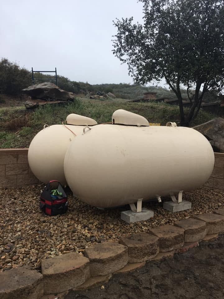 Three cream-colored cylindrical tanks on gravel beside a stone wall in a rural yard with trees.