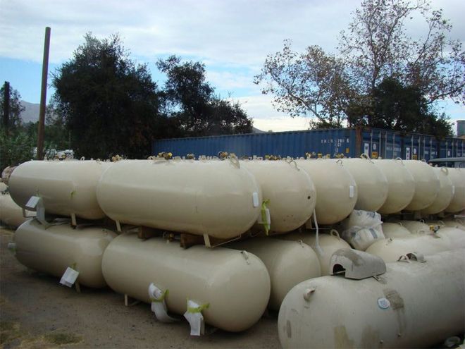 Stacked beige propane tanks outdoors beside a blue fence under a cloudy sky