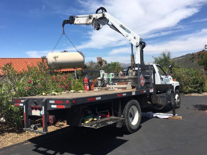 Flatbed truck with a crane lifting a white tank in a sunny driveway or worksite