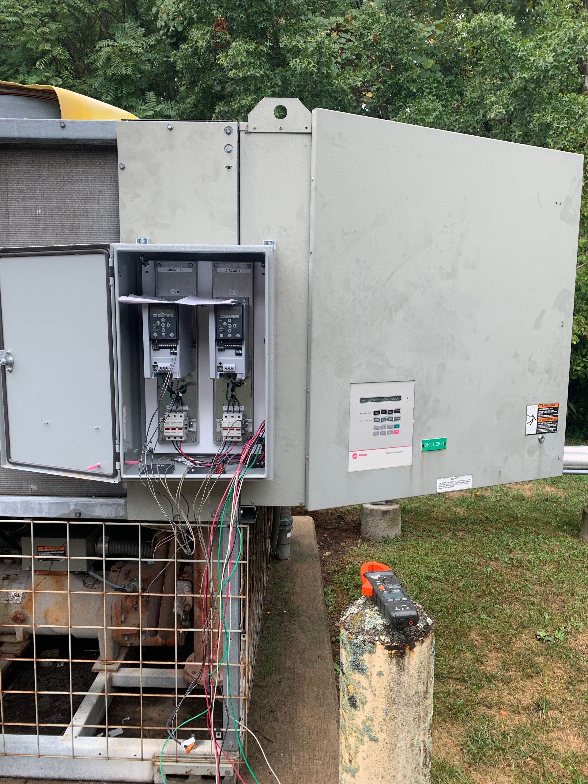 A large electrical box is sitting in the grass next to a fence.