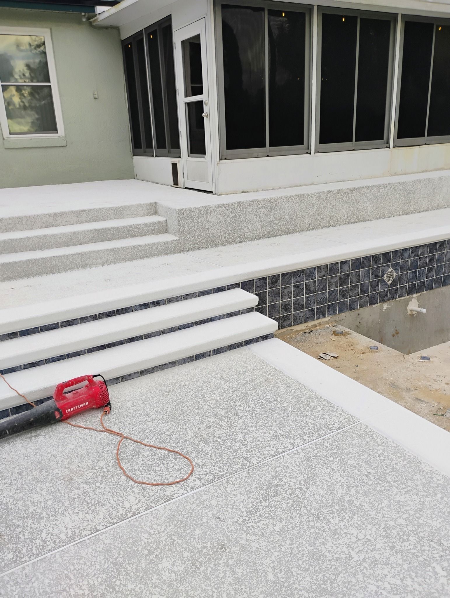 Outdoor patio with speckled concrete, steps, and large windows; construction tools and pipes in the foreground
