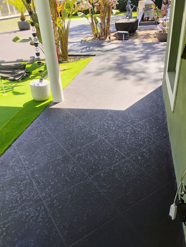 Covered patio with black tiled floor, green artificial turf, and potted plants in a sunny garden.