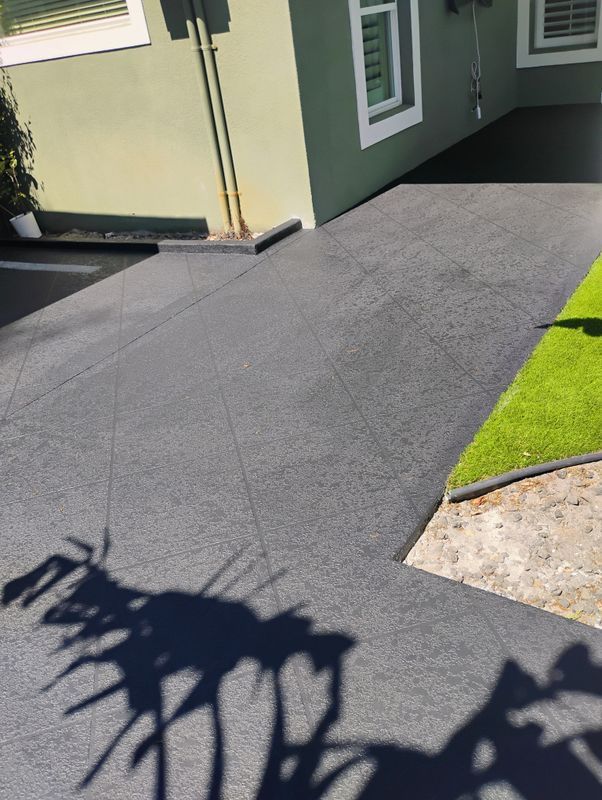 Freshly paved dark driveway beside a green house, with a small grass patch and tree shadows in the foreground
