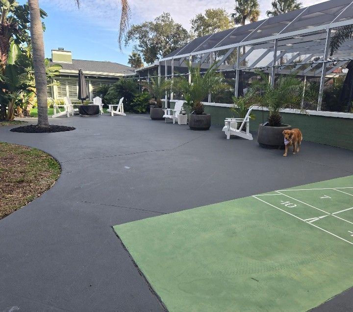 Outdoor patio with black pavement, green turf, white chairs, potted plants, and a metal pergola.