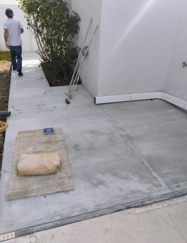 Concrete side yard with a person walking away, a small tree, and a utility cover on the ground.