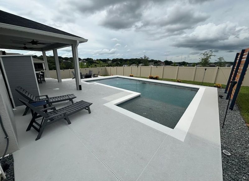 Modern backyard pool with white patio, lounge chairs, and cloudy sky.