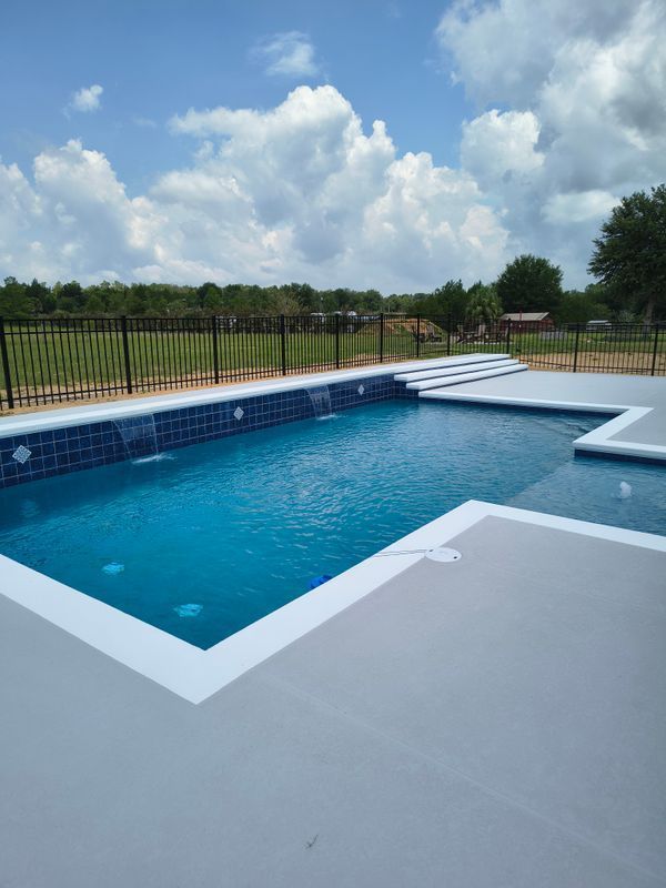Rectangular backyard pool with blue water, white deck, and black fence under a cloudy sky.