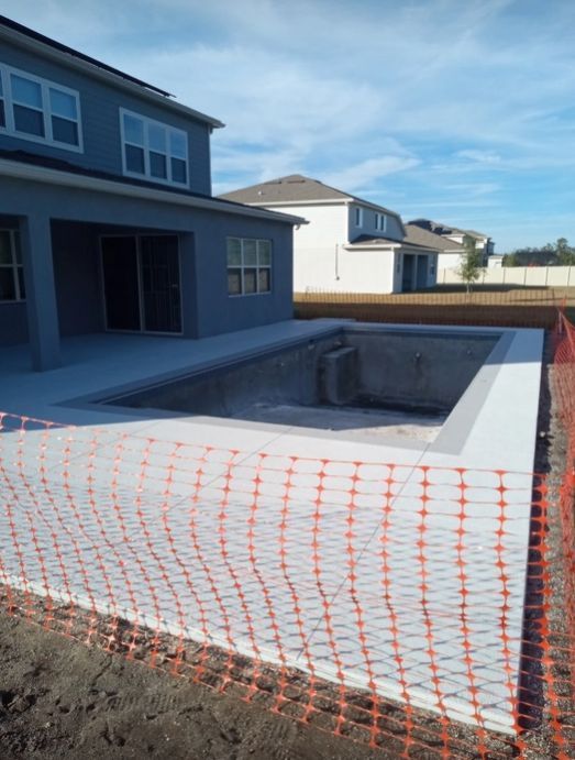 Backyard concrete pool under construction, surrounded by orange safety netting beside a house