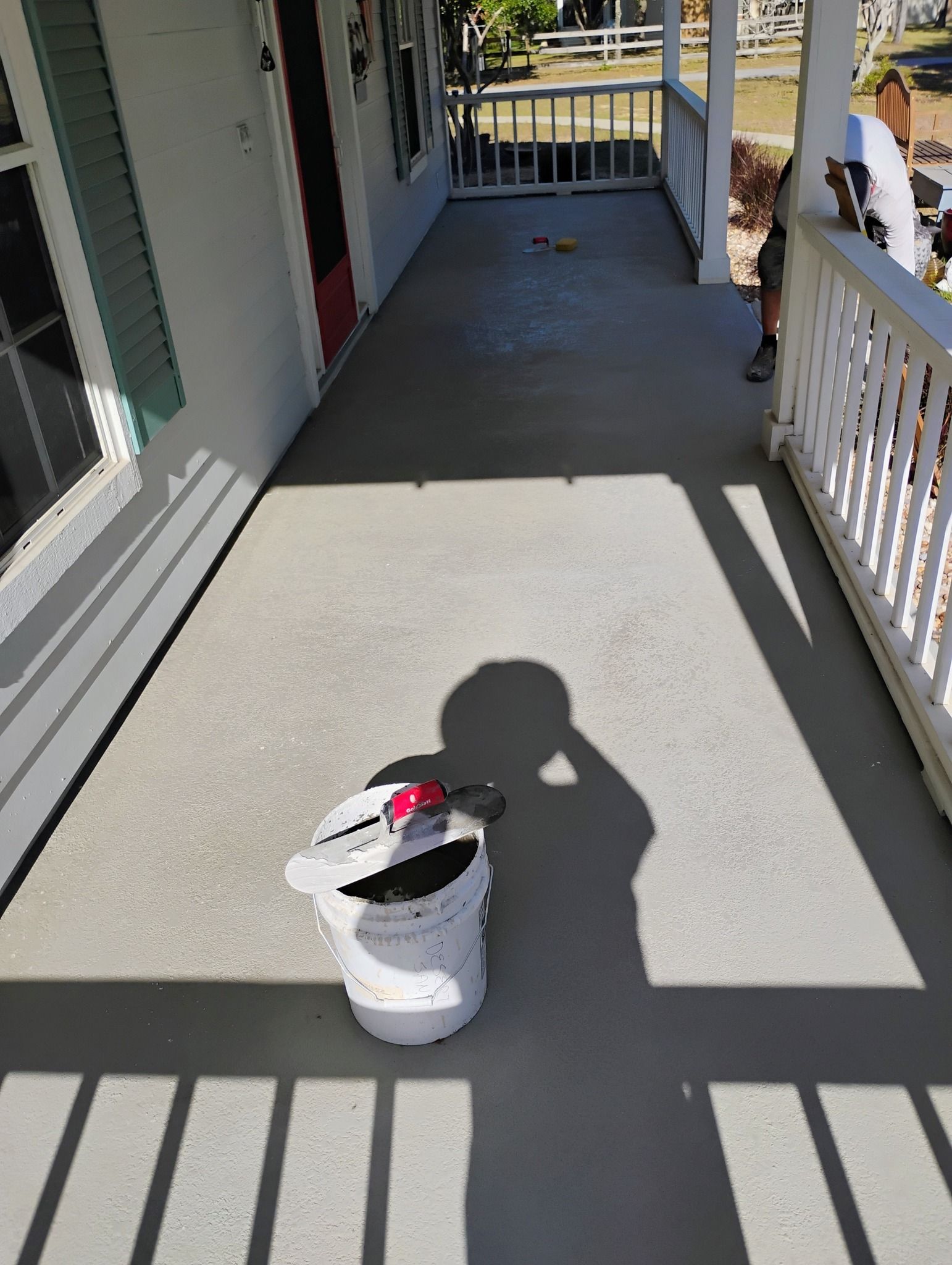 White bucket with tools on a sunlit porch, with railing shadows and house siding visible.