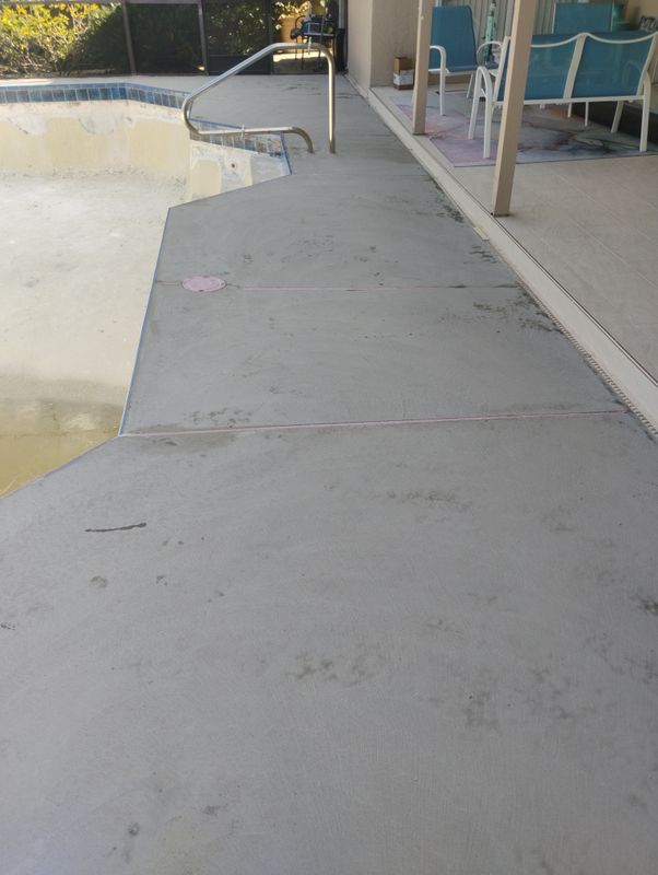 Empty concrete pool deck beside a yellow pool wall, with chairs and a handrail in the background