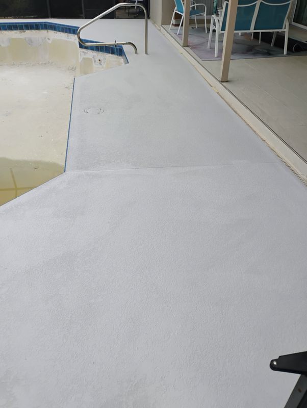 White concrete pool deck beside a swimming pool, with a blue handrail and surrounding patio seating.