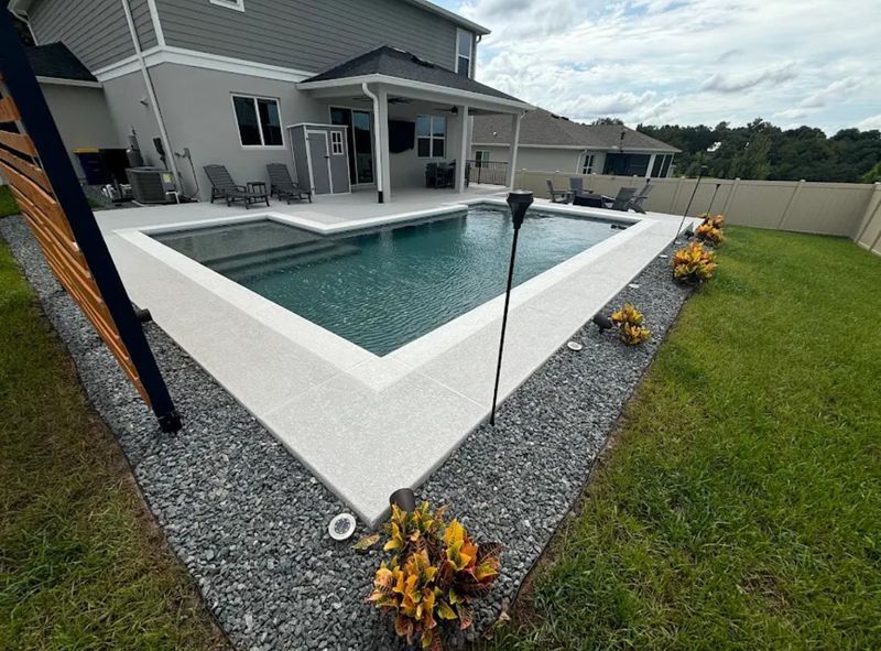 Modern backyard with a rectangular in-ground pool, white coping, gray gravel border, and lawn beside a house.