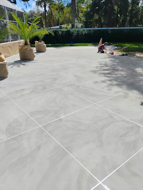 Sunny patio with light gray concrete tiles, potted palms, and a person relaxing near the edge of a lawn.