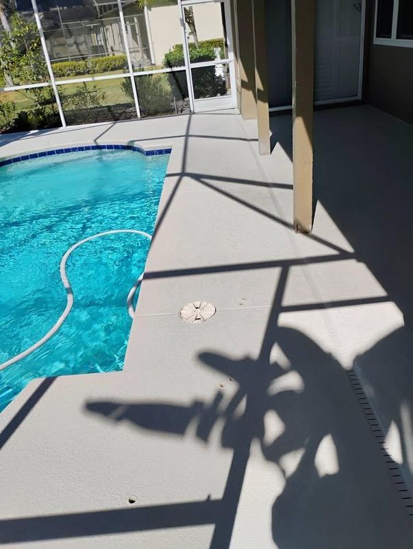 Poolside patio with bright blue water, gray concrete walkway, and shadows from a nearby building and railing.