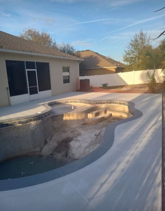 Empty backyard with a damaged, dry swimming pool next to a house and patio.